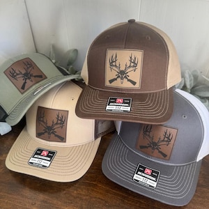 May include: A collection of Richardson 112 adjustable trucker hats in various colors, including brown, tan, gray, and olive green. Each hat features a leather patch with a deer antler and rifle design. The hats have a mesh back and a curved brim.