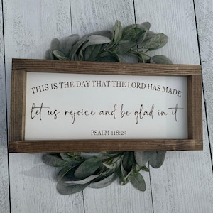 May include: A rectangular wooden sign with the text "THIS IS THE DAY THAT THE LORD HAS MADE, let us rejoice and be glad in it, PSALM 118:24" in black script. The sign is framed in dark brown wood and surrounded by a green leafy wreath.