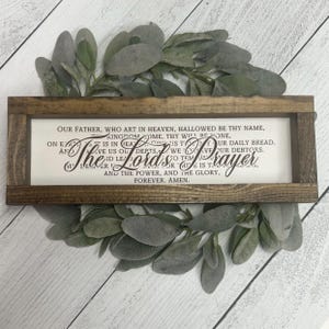 May include: A wooden sign with a white background and brown lettering. The sign features the Lord's Prayer, a traditional Christian prayer. The sign is perfect for adding a touch of faith to any home.