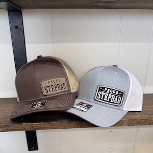 May include: Two baseball caps with the text "PROUD STEPDAD" on a rectangular patch. One cap is brown with a tan mesh back, the other is gray with a white mesh back. Both hats are on a wooden shelf.