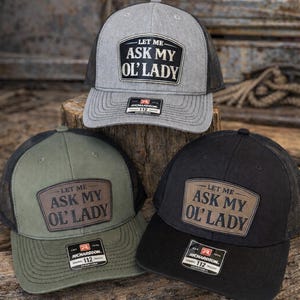 May include: Three trucker hats in gray, olive green, and black, each with a mesh back and a curved brim. The hats feature a rectangular patch with the text "LET ME ASK MY OL' LADY". The Richardson 112 trucker hat is adjustable.