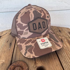 May include: A brown camouflage baseball cap with a mesh back. The front of the cap features a black hexagon with the word "DAD" in bold letters. A white tag with black text is attached to the brim.