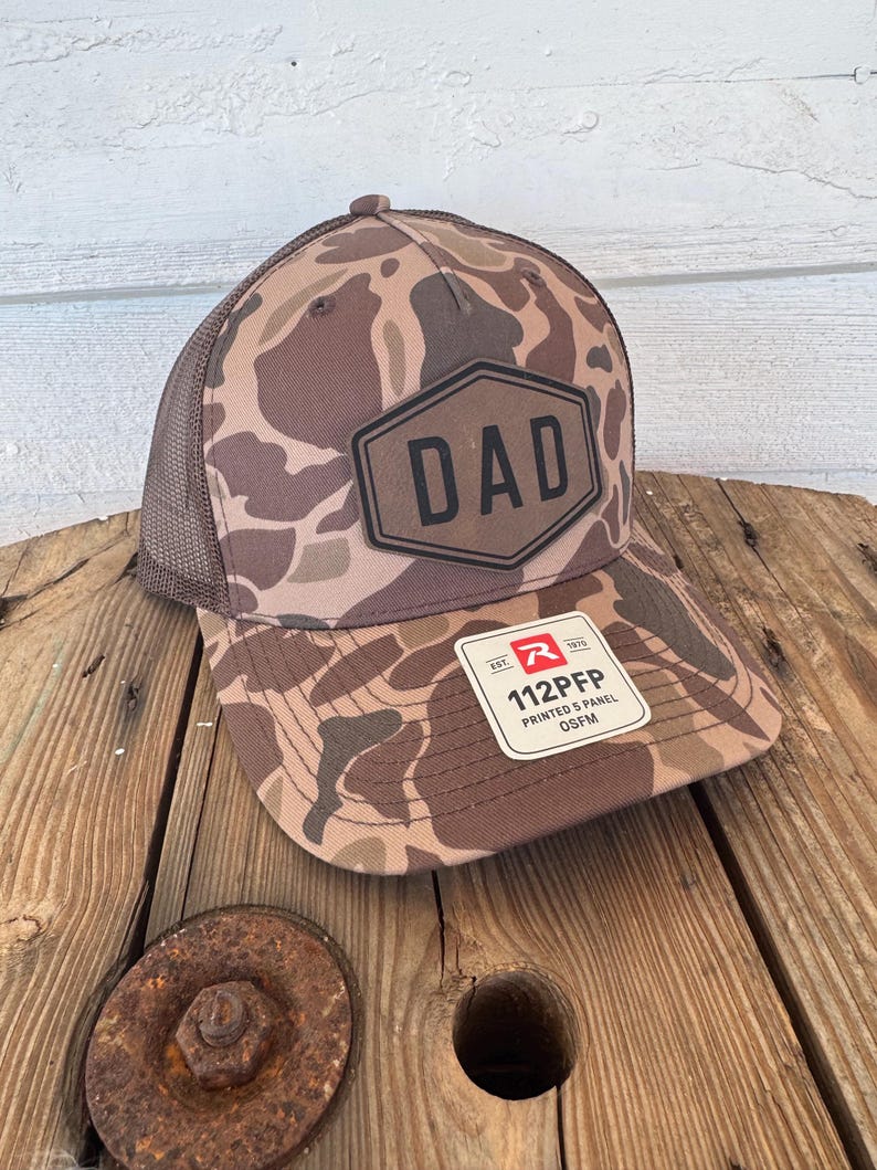 May include: A brown camouflage baseball cap with a mesh back. The front of the cap features a black hexagon with the word "DAD" in bold letters. A white tag with black text is attached to the brim.
