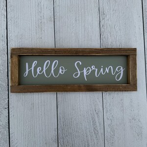 May include: A rectangular wooden sign with the words "Hello Spring" in white script lettering. The sign has a sage green background and a dark brown wooden frame. The sign is displayed against a white wooden plank background.