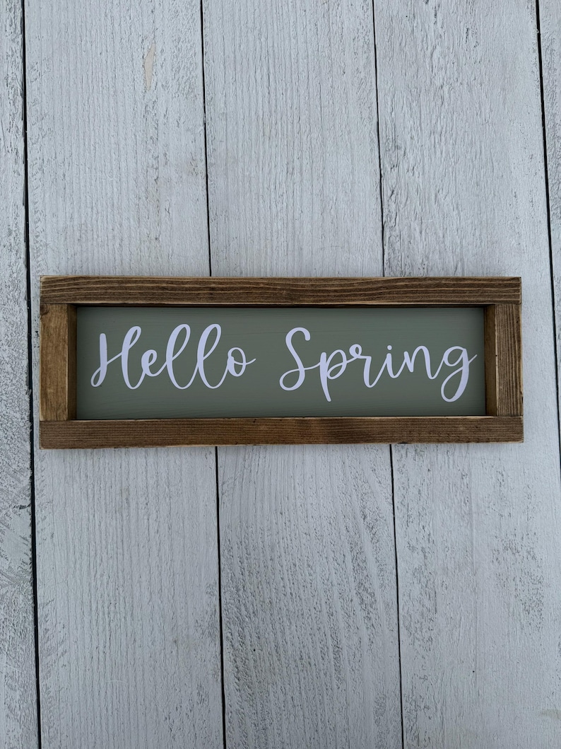 May include: A rectangular wooden sign with the words "Hello Spring" in white script lettering. The sign has a sage green background and a dark brown wooden frame. The sign is displayed against a white wooden plank background.