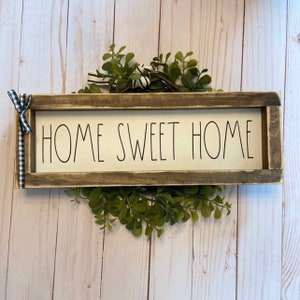 May include: A rustic wooden sign with the text "HOME SWEET HOME" in black lettering on a cream background. The sign is framed with a dark brown wood frame and has a black and white checked ribbon tied to the top.