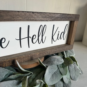 May include: A wooden sign with a white background and a brown frame. The sign says "The Hell Kid".