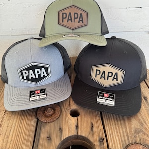 May include: Three trucker hats with the word "PAPA" on a leather patch. The hats are in olive green, gray, and black, each with a mesh back. The hats are displayed on a wooden surface.