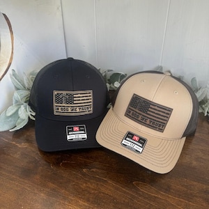 May include: Two trucker hats with a black mesh back. One hat is black with a brown leather patch that says "In God We Trust" and an American flag. The other hat is tan with a brown leather patch that says "In God We Trust" and an American flag. Both hats have a Richardson 112 label.