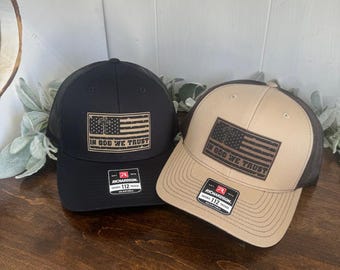 Puede incluir: Dos gorras de camionero con una parte trasera de malla negra. Una gorra es negra con un parche de cuero marrón que dice "In God We Trust" y una bandera estadounidense. La otra gorra es de color beige con un parche de cuero marrón que dice "In God We Trust" y una bandera estadounidense. Ambas gorras tienen una etiqueta Richardson 112.