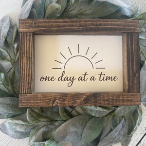 May include: A small, rectangular wooden sign with a light background and a dark brown frame. The sign features a sun graphic and the phrase "one day at a time" in a handwritten style font. The sign is surrounded by a green and gray leafy wreath.