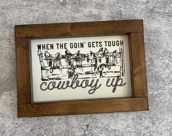 May include: A wooden framed sign with a white background and brown text that reads "When the goin' gets tough, cowboy up". The sign features a black and white illustration of saddles hanging on a fence.