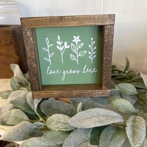 May include: A small wooden sign with a green background and white floral designs. The sign reads "love grows here".
