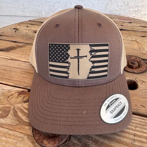 May include: A brown snapback cap with a mesh back and a rectangular patch on the front. The patch features a distressed American flag design with a black cross in the centre. A circular sticker on the brim reads The Authentic Yupoong Snapback.