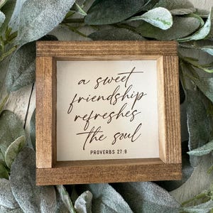 May include: A small, square wooden sign with the text "a sweet friendship refreshes the soul" and "PROVERBS 27:9" in elegant script. The sign has a light-colored background and a dark brown frame, surrounded by green and gray foliage.