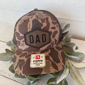 May include: A brown and tan camouflage baseball cap with a mesh back. The cap features a leather patch with the word "DAD" in bold letters. A white tag with the text "112PFP" is attached to the brim.