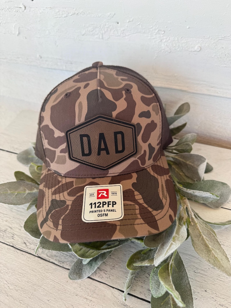 May include: A brown and tan camouflage baseball cap with a mesh back. The cap features a leather patch with the word "DAD" in bold letters. A white tag with the text "112PFP" is attached to the brim.