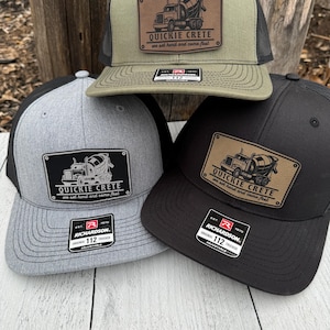 May include: Three trucker caps in olive green, grey, and black. Each cap has a rectangular patch with a cement truck graphic and the text "QUICKIE CRETE we set hard and come fast." The caps have a mesh back and a curved peak.