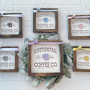 May include: Six wooden signs with a rustic brown frame and white background. Each sign has a different colored ribbon and a coffee cup graphic. The text on each sign reads "Cottontail Coffee Co. Hop on In!" with the words "Fresh Brewed" and "Self Served" above and below the coffee cup graphic.