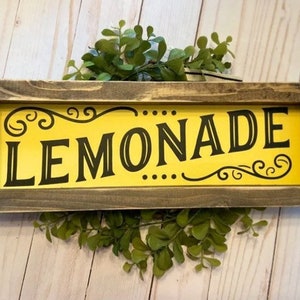 May include: A rectangular wooden sign with the word "LEMONADE" in black, stylized font on a yellow background. The sign has a dark gray frame and decorative flourishes. Greenery is placed behind the sign.