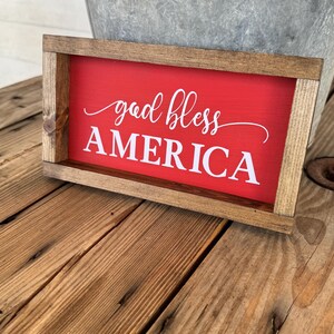 Puede incluir: Un letrero rectangular de madera con fondo rojo y las palabras "god bless AMERICA" en escritura blanca. El letrero est&aacute; enmarcado en madera natural y descansa sobre una superficie de madera.