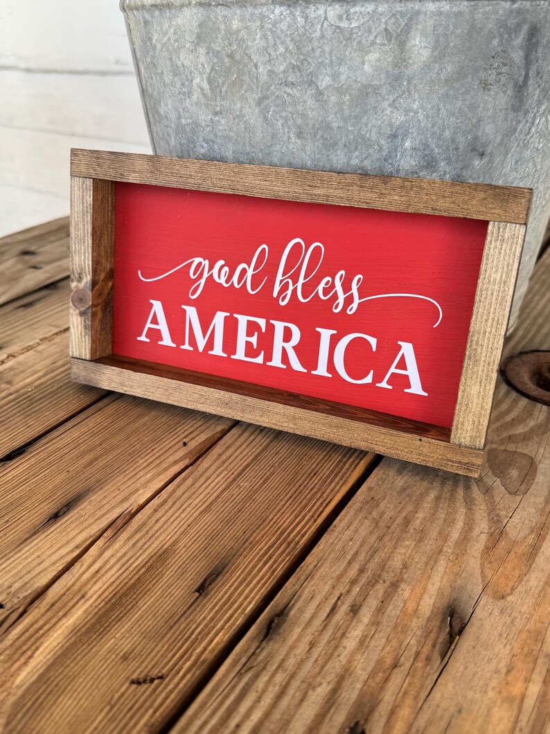 Puede incluir: Un letrero rectangular de madera con fondo rojo y las palabras "god bless AMERICA" en escritura blanca. El letrero est&aacute; enmarcado en madera natural y descansa sobre una superficie de madera.