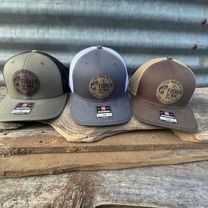May include: Three trucker hats with a curved brim are displayed on a wooden surface. Each hat has a different color scheme: olive green, gray, and brown. The hats feature a circular patch with the text "JESUS MADE ME A FISHER OF MAN, I CAN CATCH 'EM CLEAN."