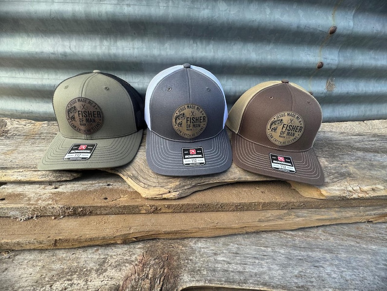 May include: Three trucker hats with a curved brim are displayed on a wooden surface. Each hat has a different color scheme: olive green, gray, and brown. The hats feature a circular patch with the text "JESUS MADE ME A FISHER OF MAN, I CAN CATCH 'EM CLEAN."