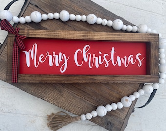 Puede incluir: Un letrero de madera con fondo rojo y letras blancas que dice "Feliz Navidad". El letrero está enmarcado en madera marrón oscura y tiene una cinta roja y negra a cuadros atada en la parte superior.