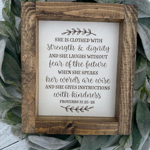 May include: A framed wooden sign with the text "SHE IS CLOTHED WITH strength & dignity" and other inspirational words from Proverbs 31:25-26. The sign has a rustic, brown frame and is surrounded by green and gray foliage.
