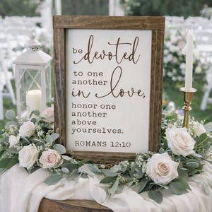 May include: A wooden framed sign with the words "be devoted in love" in elegant script. The sign is surrounded by white roses and greenery, set on a wooden barrel. The text "Romans 12:10" is at the bottom.