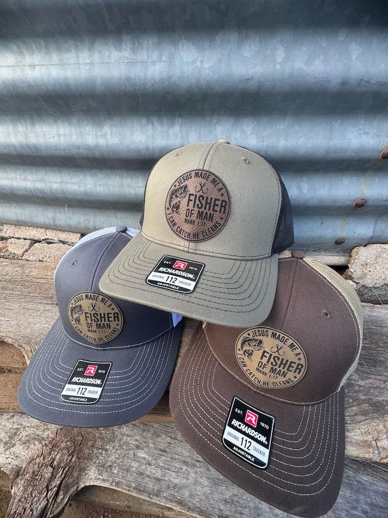 May include: Three trucker hats in shades of gray, olive green, and brown, stacked on a wooden surface. Each hat features a circular patch with the text "JESUS MADE ME A FISHER OF MAN CAN CATCH HE CLEANS". The hats are adjustable.