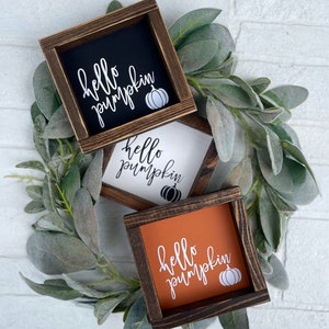May include: Three small, square wooden signs with the words "hello pumpkin" in white script and a pumpkin graphic. The backgrounds are black, white, and orange. The signs are surrounded by a green leafy wreath.