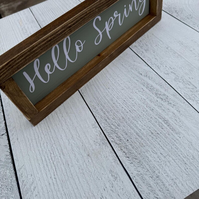 May include: A rectangular wooden sign with the words "Hello Spring" in white script on a green background. The sign is framed in dark brown wood and rests on a white-painted wooden surface.