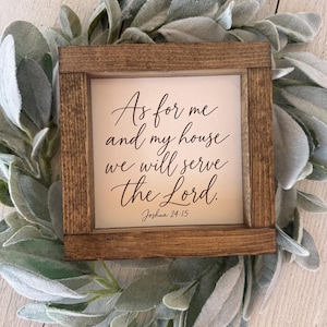 May include: A square wooden sign with a dark brown frame and a white center panel. The text reads, "As for me and my house we will serve the Lord. Joshua 24:15" in a black cursive script. The sign is surrounded by a wreath of green leaves.