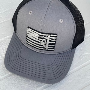 May include: Gray and black trucker hat with a curved brim. The front panel features a patch with a black and white American flag design and a silhouette of a lineman on a utility pole. The hat has a mesh back.