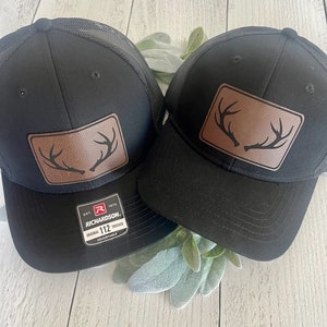 Peut inclure: Deux casquettes de camionneur en maille noire avec des patchs en cuir marron représentant une silhouette de bois de cerf. La casquette de gauche porte le texte "EST. 1970 RICHARDSON. ORIGINAL 112 TRUCKER ADJUSTABLE" sur le patch.