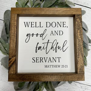 May include: A framed wooden sign with the text "WELL DONE, good AND faithful SERVANT" and the Bible verse Matthew 25:21. The sign has a white background and is surrounded by a brown wooden frame. The sign is set on a bed of green leaves.