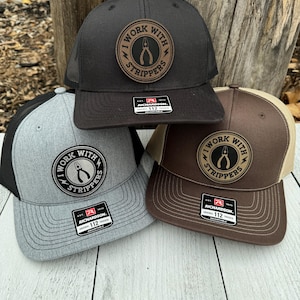 May include: Three trucker hats in black, gray, and brown, each featuring a circular patch with the text "I WORK WITH STRIPPERS" and a pliers graphic. The hats have mesh backs and adjustable closures.