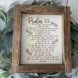 May include: A wooden framed sign featuring the text of Psalm 23 on a cream background. The frame is dark brown, and the sign is surrounded by green foliage. The text is in black.