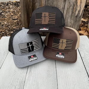 May include: Three baseball caps with a mesh back and a flat brim. Each cap features a patch with an American flag design and a cross. The caps are in black, grey, and brown with a tan or black mesh.