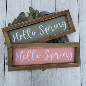 May include: Two rectangular wooden signs with the words "Hello Spring" in white script. One sign has a sage green background, the other a soft pink. The signs are framed in a dark brown wood.