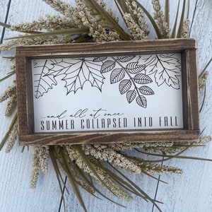 May include: A wooden sign with a white background and black lettering. The sign features a design of leaves and the text "...and all at once SUMMER COLLAPSED INTO FALL".