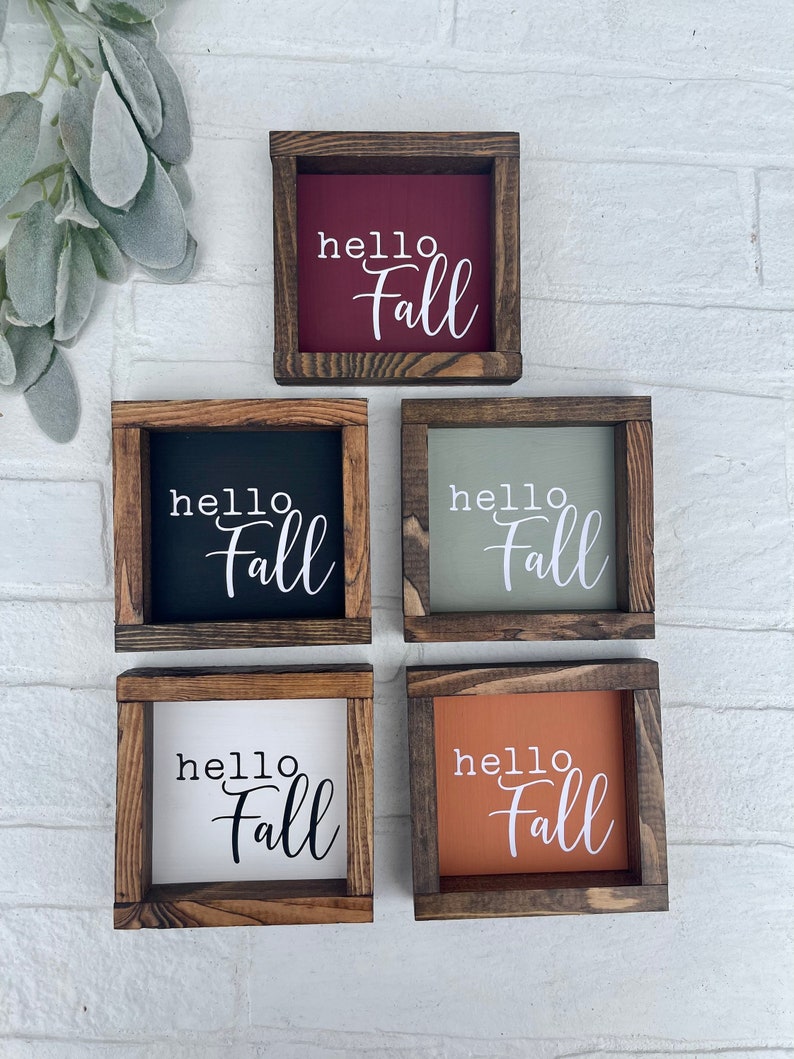 May include: Five wooden signs with the text 'hello fall' in white lettering. The signs are in different colors: burgundy, black, green, white, and orange. The signs are all the same size and shape and have a rustic wood frame.