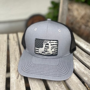 May include: Gray trucker hat with a black mesh back and a curved brim. The front features a rectangular patch with an American flag design and a silver excavator silhouette. The hat is sitting on a wooden surface.