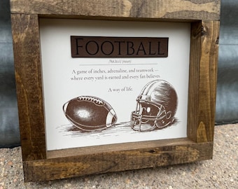 May include: Wooden framed sign with the word "FOOTBALL" in brown lettering. The sign includes a football, a helmet, and text that reads "A game of inches, adrenaline, and teamwork - where every yard is earned and every fan believes."
