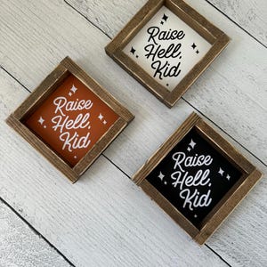 May include: Three square wooden signs with the phrase "Raise Hell, Kid" in white script. The signs have different coloured backgrounds: white, orange, and black. Each sign is framed in a dark brown wood.