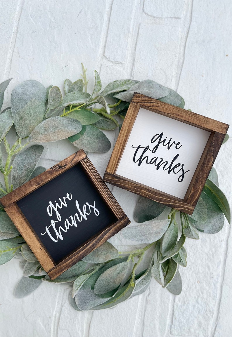 May include: Two wooden signs with the text "give thanks" in white lettering. One sign has a black background and the other has a white background. The signs are surrounded by a green leafy garland.