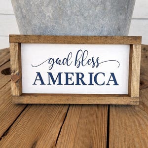 Puede incluir: Un letrero rectangular de madera con las palabras "god bless AMERICA" en letras azul marino. El letrero tiene un fondo blanco y un marco de madera marr&oacute;n. El letrero se muestra sobre una superficie de madera.