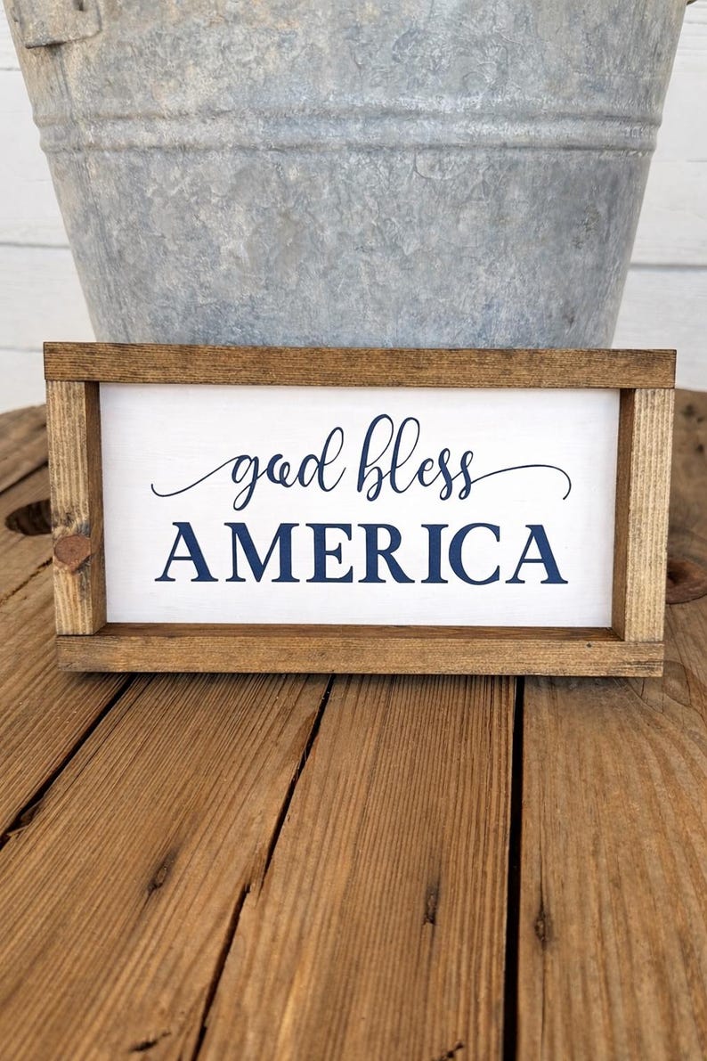 Puede incluir: Un letrero rectangular de madera con las palabras "god bless AMERICA" en letras azul marino. El letrero tiene un fondo blanco y un marco de madera marr&oacute;n. El letrero se muestra sobre una superficie de madera.